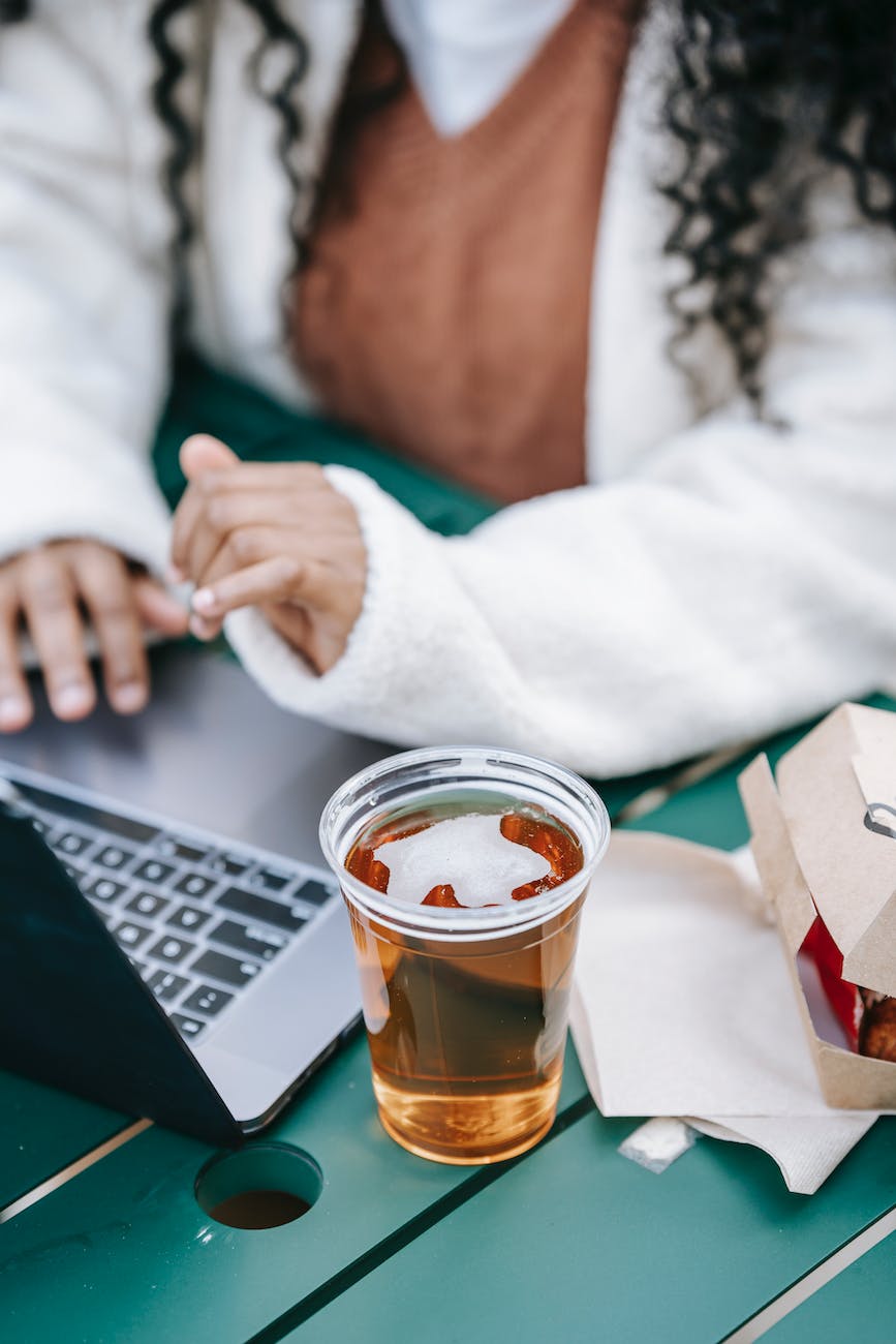 crop black faceless woman with glass of beer using laptop